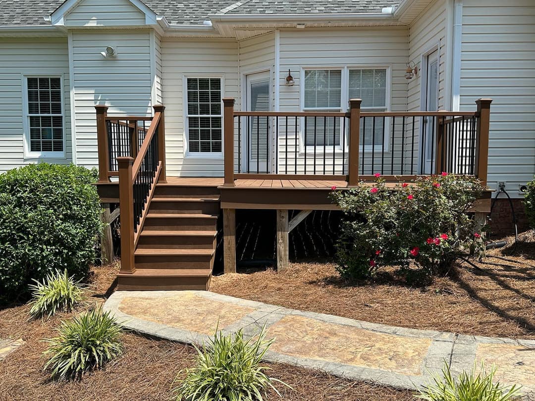 Wooden deck with railings and steps, surrounded by shrubs and a stone pathway.