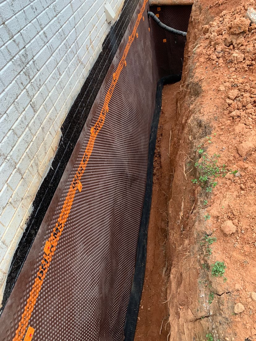 Drainage system installation in a trench beside a brick wall, showcasing waterproofing membrane.