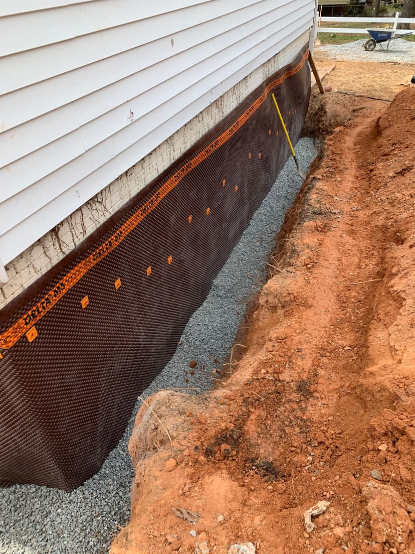 Foundation drainage system installation with gravel and drainage fabric beside a house.