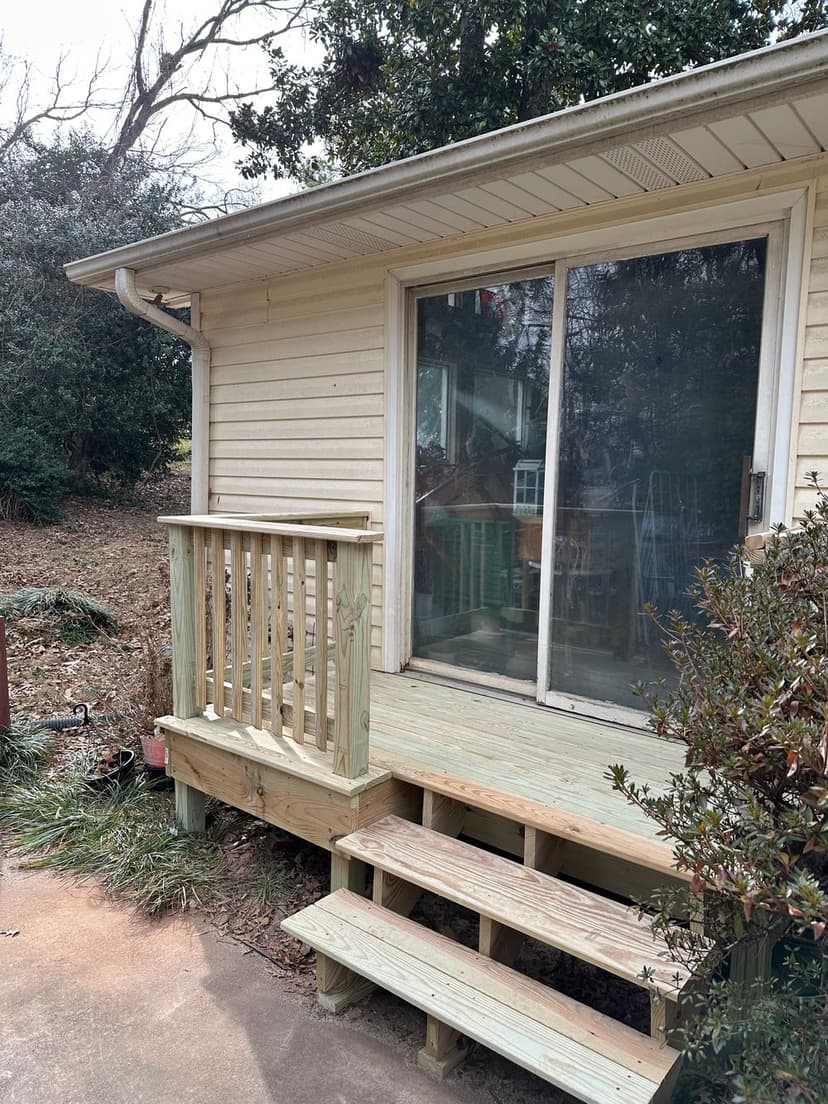 Wooden deck and steps leading to a glass sliding door of a house. Outdoor greenery visible.